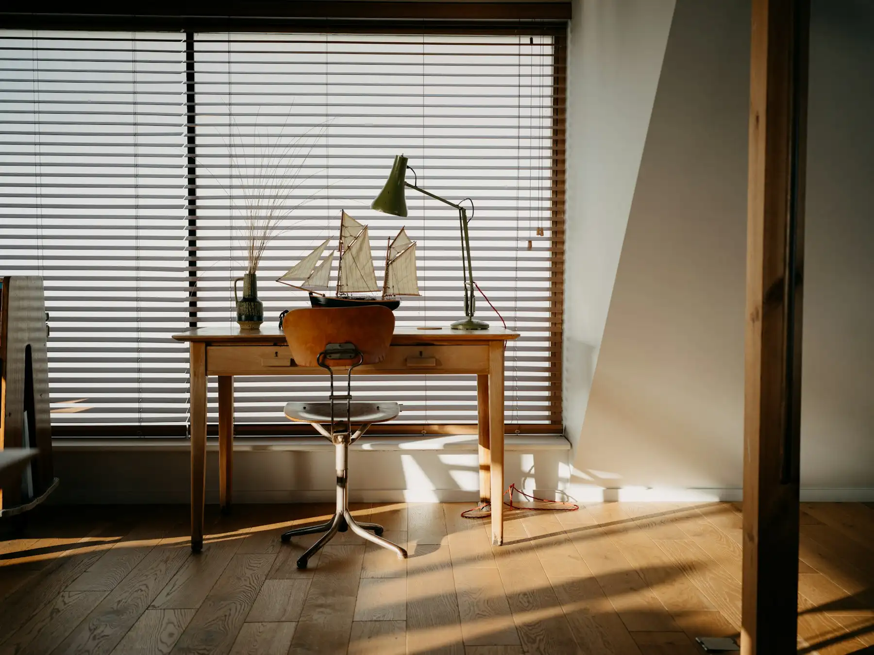 Bureau en bois massif devant une fenêtre à stores, lampe d’architecte et soleil rasant